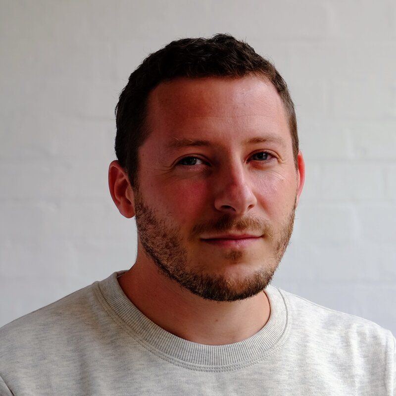 Julien Sansot, smiling slightly, in a light grey sweater against a white brick wall